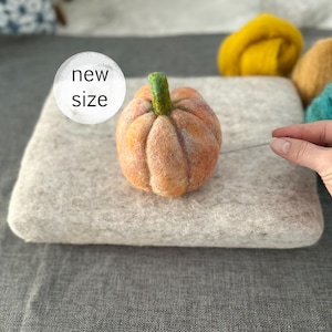 以下が含まれることがあります： オレンジと紫の色合いの手作りのフェルトカボチャ。緑色の茎があり、灰色のフェルトのベースの上に置かれています。「new size」というテキストが白い円の中にあります。秋をテーマにした手作りのアイテムです。