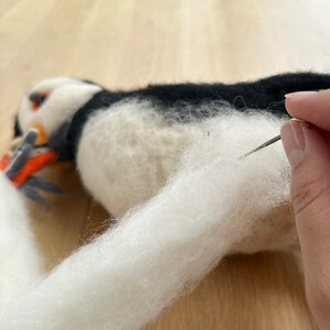 May include: A white and black needle felted puffin with a white tail being felted with a needle.