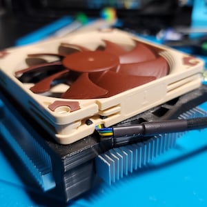 Puede incluir: Un refrigerador de CPU de computadora beige con un ventilador marrón y un cable negro. El refrigerador está sobre una superficie azul.