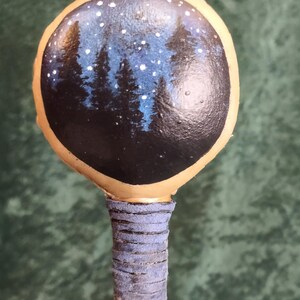 May include: A hand-painted wooden wand with a circular design of a starry night sky over a forest. The wand has a light brown handle with a blue leather grip and a decorative pattern.