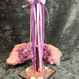 May include: A handcrafted wand with a dark brown wooden shaft, adorned with a floral crown of pink and green roses, and flowing purple and white ribbons. The wand rests on a square base decorated with small stones and faux greenery.