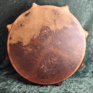 May include: A circular frame drum featuring a stretched, natural tan and dark brown hide head. The drum head displays a mottled, organic texture with visible grain patterns and is secured around the edges by folded leather lacing points.
