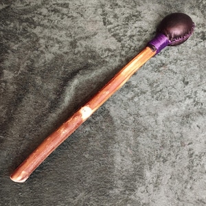 May include: A percussion mallet featuring a long, cylindrical wooden handle with a natural brown grain. The head is a rounded, dark brown leather-covered sphere, securely attached to the handle with a vibrant purple thread wrapping.