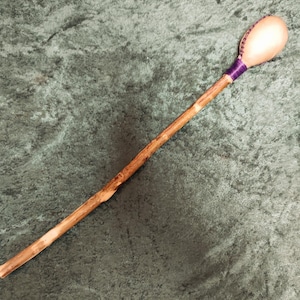 May include: A handcrafted wooden rattle with a light brown handle and a rounded, light-coloured head. The head is wrapped with purple thread and has a patterned design. The rattle is set against a green background.