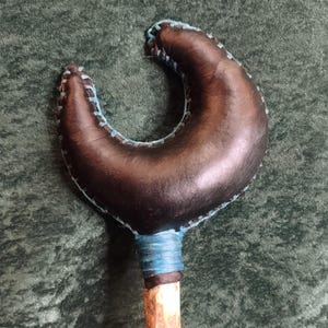 May include: A crescent-shaped object made of dark brown leather with light blue stitching along the edges. The crescent is attached to a light-coloured wooden handle, with the connection point wrapped in blue material for a secure finish.
