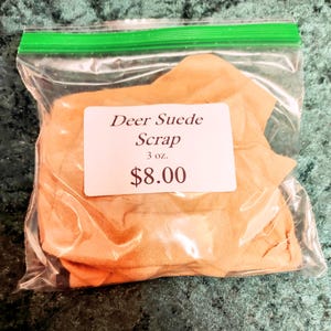 May include: A clear plastic bag containing deer suede scraps. A white label displays the text "Deer Suede Scrap" and the weight, 3 oz. The price is $8.00. The suede is a light tan color.