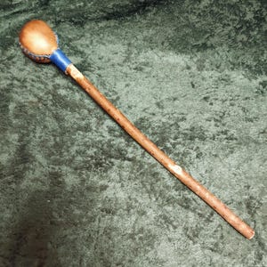 May include: A handcrafted wooden staff with a rounded, light brown head. The head is attached to the staff with blue leather and stitching. The staff is made of a natural, light brown wood.