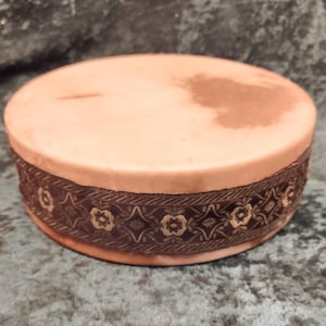 May include: A round, light peach-colored container with a smooth, flat top surface. The side of the container is wrapped in a dark brown decorative ribbon featuring a repeating pattern of small, light-colored floral and geometric shapes.