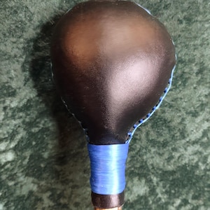 May include: A percussion instrument with a smooth, dark brown rounded head and a textured wooden handle. The head is secured to the handle with bright blue lacing and a matching blue wrapped grip section, creating a distinct contrast in colour.