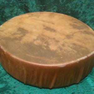 Pode incluir: Um tambor redondo de topo plano com uma pele bronzeada e manchada e uma estrutura de madeira castanha texturizada. O tambor apresenta uma superfície de jogo circular lisa com variações de cor naturais e uma parede lateral cilíndrica.