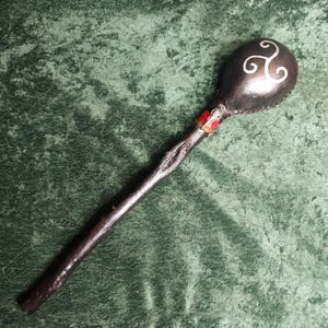 May include: A black rattle with a long, textured handle and a rounded head featuring a white triskelion symbol. The neck of the rattle is decorated with a band of red and silver-toned metal accents, and the head is stitched along the side.