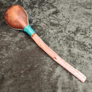 May include: A percussion instrument featuring a rounded, brown leather-covered head stitched with light blue thread. The head is attached to a light-coloured, textured wooden handle wrapped with a vibrant turquoise band at the connection point.