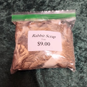 May include: A clear plastic bag filled with rabbit fur scraps. A white label on the bag reads "Rabbit Scrap" with the weight of 3 oz. The bag is sealed with a green zipper closure. The background is a dark green fabric.
