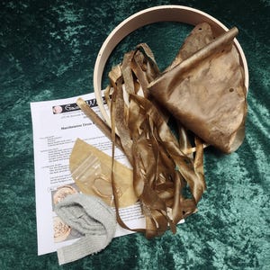 May include: A DIY drum kit with instructions, a wooden hoop, a piece of leather, a stick, a grey cloth, and a bag of twine. The kit is laid out on a green velvet surface.
