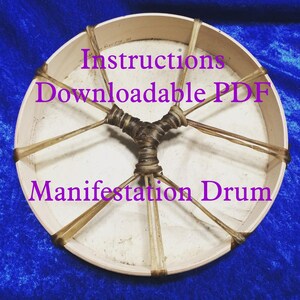 May include: A white wooden frame drum with brown leather lacing. The drum is surrounded by a blue fabric background. The text "Instructions Downloadable PDF" and "Manifestation Drum" is printed in purple on the drum.