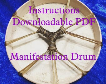 Può includere: Un tamburo a cornice in legno bianco con lacci in pelle marrone. Il tamburo è circondato da uno sfondo in tessuto blu. Il testo "Instructions Downloadable PDF" e "Manifestation Drum" è stampato in viola sul tamburo.