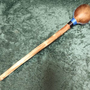 May include: A handcrafted percussion instrument, a maraca, with a wooden handle and a rounded head. The head is covered in brown leather, accented with blue and black bands. The maraca is designed for musical performance.