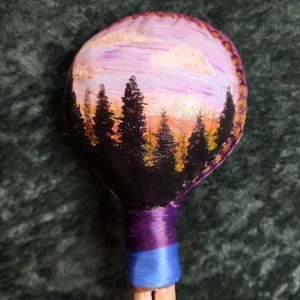May include: A round, hand-painted maraca featuring a sunset landscape with black silhouettes of pine trees against a purple, orange, and yellow sky. The handle is wrapped in purple and blue thread, and the top edge is stitched with brown cord.