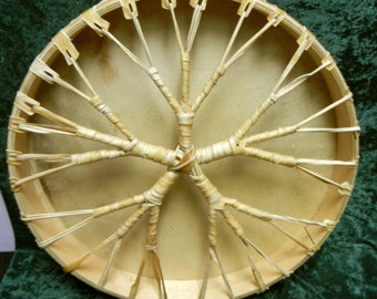 May include: A close-up of the back of a round, wooden frame drum with a natural leather head. The drum is held together with a woven pattern of natural fibres.