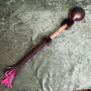 May include: A long, slender staff featuring a dark brown leather-covered handle, a central wooden section, and a rounded top end stitched with bright pink thread. The handle is accented with dark fur trim and ends in a fringe of pink and brown strips.