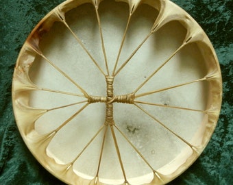 May include: A round, tan-coloured Native American drum with a cross-shaped lacing pattern and 12 spokes radiating from the centre.