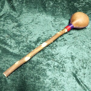 May include: A handcrafted percussion instrument, possibly a rattle or shaker, with a light brown leather head attached to a wooden handle. The handle is a natural branch with a light brown colour and white spots. The head is secured with red and purple bands.
