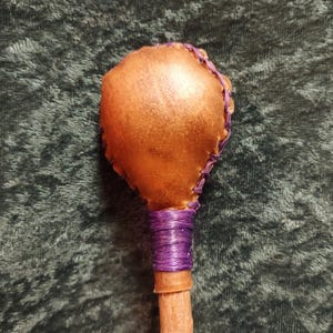 May include: A handcrafted wand with a brown leather head, stitched with purple thread. The handle is wrapped in purple thread and attached to a wooden dowel. The wand is set against a dark green background.