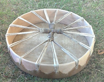 May include: A circular drum with a natural hide drumhead and a light-coloured wooden frame. The drum is laced with light-coloured leather straps that radiate from a central, wrapped handle. The drum is resting on green grass.