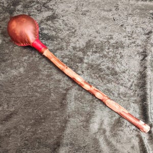 May include: A long wooden staff featuring a rounded, brown leather-covered head at the top. The leather head is stitched with red thread and secured to the wooden handle with a bright red wrapped band. The handle is a natural, textured branch.