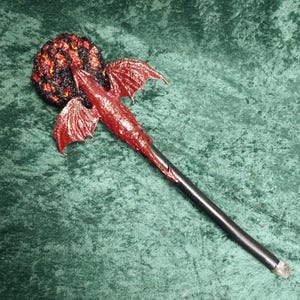 May include: A fantasy staff with a red dragon design. The staff has a black handle, a red dragon body with wings, and a textured, fiery orb at the top. The dragon's wings are spread, and the orb has orange and yellow accents.