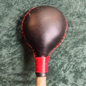 May include: A black leather-covered round head attached to a wooden handle. The leather head features prominent red stitching along the outer edge and a thick red cord wrapped tightly around the neck where the head meets the wooden handle.