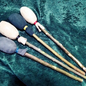 Pode incluir: Quatro baquetas de percussão com cabos de madeira e cabeças ovais. Duas cabeças são cobertas com tecido cinza escuro e duas com tecido bege claro. Cada cabeça é fixada ao cabo com um elástico colorido nas cores azul, preto, amarelo ou vermelho.