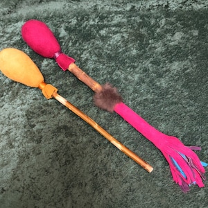 May include: Two wooden sticks with fabric covered ends. One stick has a pink fabric end with blue and purple fringe. The other stick has an orange fabric end.