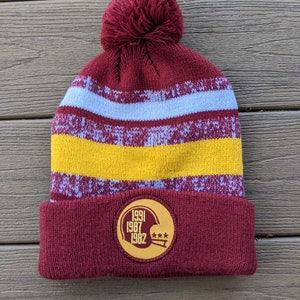 May include: A burgundy knit beanie with a white and yellow striped pattern and a pom pom on top. The beanie has a circular patch on the front with the years 1991, 1987, and 1982 printed on it, along with a football helmet graphic and three stars.