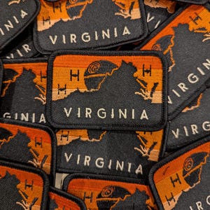May include: A collection of black and orange embroidered patches with the state of Virginia outlined in black and the word "VIRGINIA" in white text. The patches have a sunset design with an orange, peach, and beige color gradient.