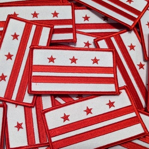 May include: A stack of red and white fabric patches featuring the flag of Washington, D.C. The patches are made of a soft, woven fabric and have a white background with three red horizontal stripes and three red stars in the upper left corner.