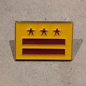 May include: A rectangular pin featuring a yellow background with three gold stars at the top and two horizontal red stripes. The pin has a gold border and is set against a gray surface.