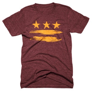 May include: A maroon t-shirt with a gold brushstroke design and three gold stars. The design is reminiscent of the Washington D.C. flag.
