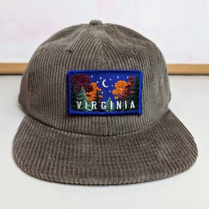 May include: A corduroy baseball cap in a muted brown color. The cap features a rectangular patch with a scenic design of trees, a crescent moon, and the word "VIRGINIA" in white lettering.