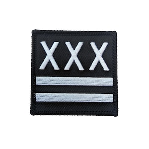 May include: Black square patch with white embroidered text and stripes. The top line features three white "X" characters. Below are two horizontal white stripes. The patch has a black border.