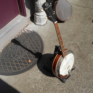 May include: A brown and white banjo with a wooden neck and a metal tuning peg. The banjo is on a stand and is in front of a door.