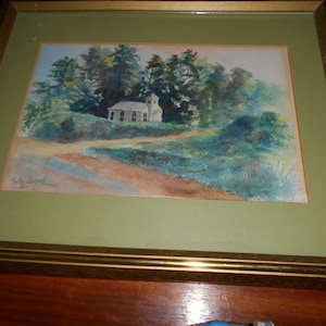 May include: Watercolor painting of a white church nestled among green trees, with a dirt road leading to it. The artwork is framed in gold and set against a green mat.
