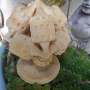 May include: A cream-colored, carved sculpture with a cluster of rounded shapes and cylindrical elements. The sculpture is mounted on a circular base with the text "RPM" and "MANUAL FOCUS". The sculpture is set in a blue pot with green plants and a statue in the background.