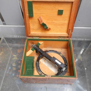May include: A vintage surveying instrument with a black metal telescope and a circular base, housed inside a square wooden storage box. The interior of the box features green felt lining, wooden support blocks, and a hinged lid with metal hardware.