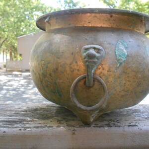 May include: A round, weathered brass pot featuring a wide rim and a bulbous body. The exterior displays a mottled patina with green oxidation spots. A decorative lion head mount is attached to the side, holding a circular metal ring handle.