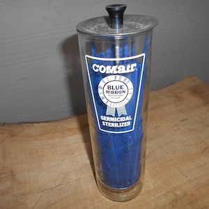 May include: A clear glass Comair Blue Ribbon Germicidal Sterilizer jar with a metal lid and black handle. The jar is filled with blue combs. The label features the text "Comair", "Blue Ribbon", and "Germicidal Sterilizer".