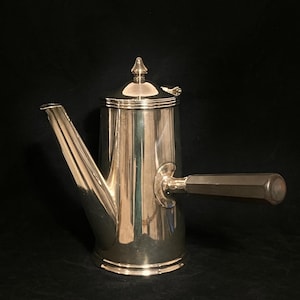 May include: A silver-colored coffee pot with a dark brown handle and a pointed lid. The pot has a long spout and a cylindrical body. The handle is attached to the side of the pot. The pot is sitting on a dark surface.