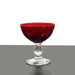 May include: A ruby red glass goblet with a round bowl and a clear, faceted stem. The goblet sits on a clear, circular base. The stem features a decorative, spherical design. The glass is likely vintage and suitable for serving beverages.