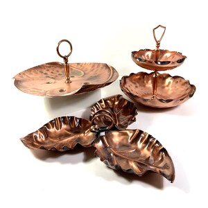 May include: A collection of copper serving dishes and trays. The set includes a two-tiered tray with a handle, a single-tier tray with a handle, and leaf-shaped dishes. The copper has a hammered texture and a warm, reddish-brown color.