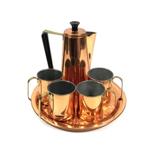May include: A vintage copper coffee set with a matching tray. The set includes a coffee pot with a black handle and lid, four mugs with gold handles, and a round tray. The copper has a shiny finish.
